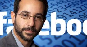 A man with glasses and a beard is standing in front of a facebook logo.